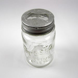 May include: A clear glass Presto Mason jar with a silver metal lid. The jar has embossed text that reads "Presto Supreme Mason". The lid has a built-in bottle opener.