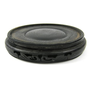 May include: A dark stained wooden display stand with a circular top and carved decorative elements. The stand is designed to hold a small object and has a low profile.