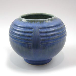 Puede incluir: Un jarrón redondo de cerámica azul con ranuras horizontales y un acento vertical. El jarrón tiene una abertura ancha y una base pequeña y redondeada. El color es azul apagado con acentos azul oscuro y verde.