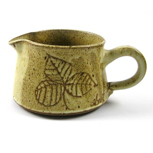May include: A small, light brown ceramic creamer with a leaf design and a brown handle. The creamer has a speckled glaze and a signature on the bottom.