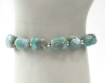 May include: A beaded bracelet featuring irregular, light blue-green stones interspersed with small silver beads. The bracelet is displayed on a white surface, showcasing the natural color variations of the stones.