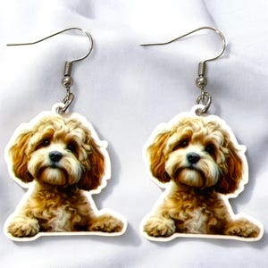 May include: A pair of dangle earrings featuring a detailed illustration of a fluffy, light brown and cream-colored dog. The earrings have silver-colored fish hook ear wires and are set against a white background.