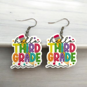 May include: A pair of white dangle earrings with the text "hello third grade" in rainbow colors. The earrings feature a pencil design and a red apple.