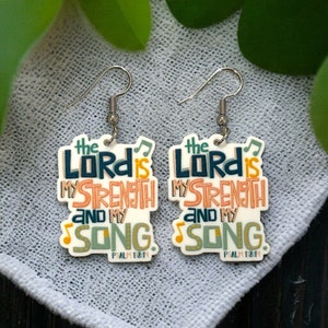 May include: A pair of white acrylic earrings with the text "The Lord is my strength and my song. Psalm 118:14" in colorful lettering.
