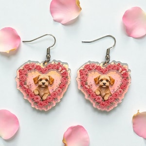 May include: A pair of heart-shaped earrings featuring a puppy illustration surrounded by pink roses and butterflies. The earrings have silver-colored hooks and are set against a white background with scattered pink rose petals.