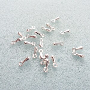May include: A group of silver-toned teardrop-shaped jewelry findings with a loop at the top. The findings are scattered on a light blue surface.