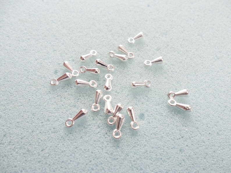 May include: A group of silver-toned teardrop-shaped jewelry findings with a loop at the top. The findings are scattered on a light blue surface.