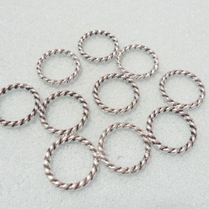 May include: A collection of silver-colored, twisted metal rings. The rings are circular and have a rope-like texture. They are scattered across a light-colored surface, suggesting a craft or jewelry-making theme.