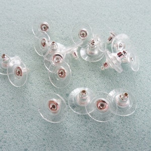 May include: A close-up of a pile of clear plastic earring backs with silver metal posts. The backs are designed to be used with stud earrings.