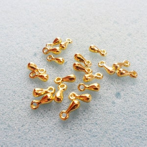 May include: A scattering of small, shiny gold-coloured jewellery findings on a light blue surface. Each finding has a teardrop-shaped body with a small loop at one end, suggesting they are designed for connecting jewellery components.