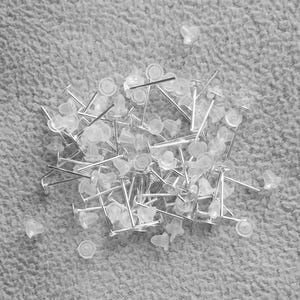 May include: A pile of silver earring posts with clear plastic backs. The posts are scattered on a white fuzzy surface.