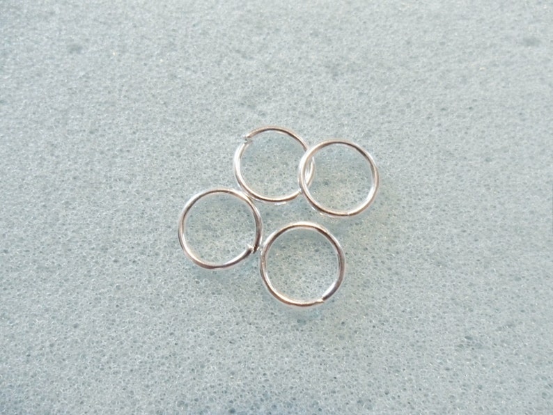 Puede incluir: Cuatro anillos de salto plateados, un tipo de hallazgo de joyer&iacute;a, est&aacute;n dispuestos sobre una superficie texturizada gris claro.