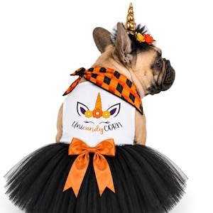May include: A white pet dress with a black tulle skirt and an orange satin bow. The bodice features a graphic of a unicorn face with the text Unicandy corn. The outfit includes an orange and black plaid bandana and a gold unicorn horn headpiece.