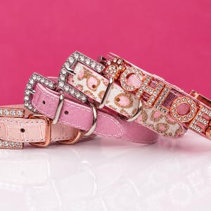 May include: Three pink pet collars with rhinestone-encrusted buckles. One collar has a light pink crocodile texture, one is solid pink, and one features a leopard print with the name CHLOE in rhinestone letters and a rhinestone paw print charm.