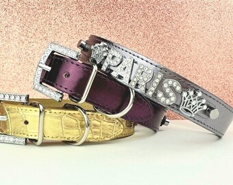 May include: Three dog collars with rhinestones. The top collar is silver with a crown and the word "PARIS" in rhinestones. The middle collar is purple and the bottom collar is gold with a crocodile pattern.