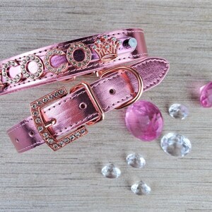 May include: Pink leather dog collar with a rose gold buckle and a crown charm. The collar is decorated with rhinestones and the word "COCCO" is spelled out in rhinestones.