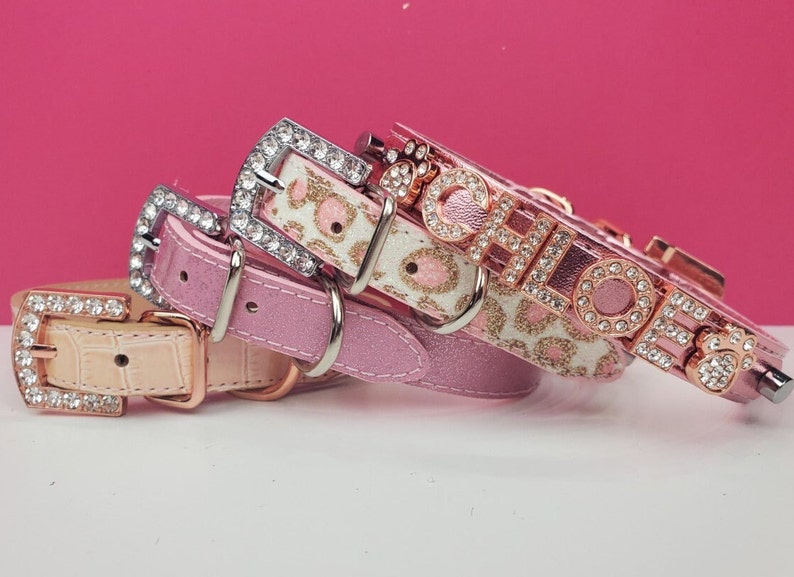 May include: Three pink dog collars with rhinestones. The top collar has the name "Chloe" spelled out in rhinestones. The middle collar has a pink and white leopard print. The bottom collar is a solid pink.