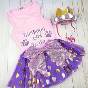 May include: Pink sleeveless shirt with the text "Birthday Girl Bella" and paw prints. A purple tulle skirt with gold polka dots and a pink sequined bow. A gold crown with white and purple flowers.