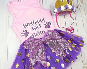 Peut inclure: Un haut rose sans manches avec le texte "Birthday Girl Bella" et des empreintes de pattes. Une jupe en tulle violette à pois dorés et un nœud en sequins rose. Une couronne dorée avec des fleurs blanches et violettes.