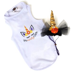 May include: A white sleeveless shirt featuring a graphic of a unicorn with a candy corn horn and the text "Unicandycorn" in black and orange. Beside it is a headpiece with a gold spiral horn, black tulle, and orange and white flowers.