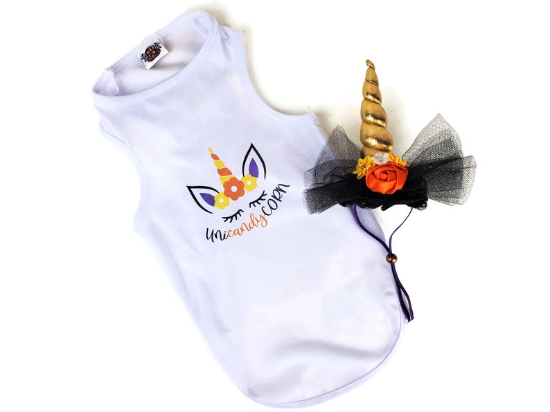 May include: A white sleeveless shirt featuring a graphic of a unicorn with a candy corn horn and the text "Unicandycorn" in black and orange. Beside it is a headpiece with a gold spiral horn, black tulle, and orange and white flowers.