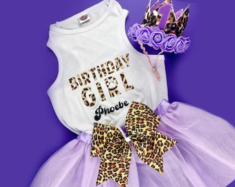 May include: A white tank top with the text "Birthday Girl" in brown and black leopard print letters. The name "Phoebe" is written below in black. A purple tulle tutu with a brown and black leopard print bow is included. A purple crown with a leopard print band and a number one is also included.