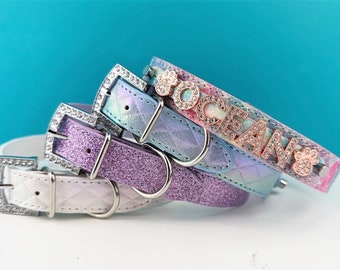 May include: Three dog collars with rhinestones and a paw print charm. The top collar is iridescent blue with the name "Ocean" in rose gold letters. The middle collar is lavender glitter and the bottom collar is white with a quilted pattern.