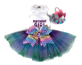 Pode incluir: Uma camiseta branca sem mangas com o texto "Birthday Girl" em glitter roxo, um laço de escamas de sereia arco-íris e uma saia de tule verde e roxo com estrelas prateadas brilhantes. Também está incluída uma coroa de feltro rosa com glitter prateado e flores de feltro turquesa e roxo.