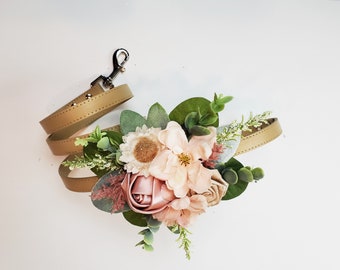 Op de afbeelding: Een bruine leren hondenriem met een zilveren sluiting, versierd met een bloemstuk van roze en witte stoffen bloemen, groene bladeren en kleine witte bloemetjes.