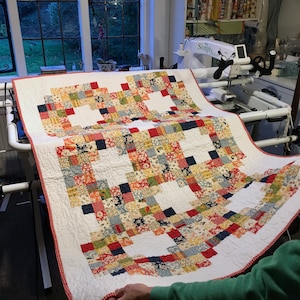 Puede incluir: Una colcha blanca con un diseño de patchwork rojo, blanco y azul. La colcha está siendo sostenida por la mano de una persona y se está exhibiendo en una máquina de acolchado de brazo largo.