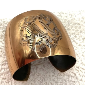May include: A wide, copper-coloured cuff bracelet with an engraved, swirling monogram design. The bracelet has a polished finish and a dark interior. The cuff is displayed on a white, textured surface.