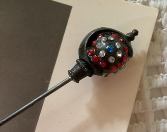 May include: A black metal pin with a round, red, white, and blue jeweled top. The pin is on a white surface.