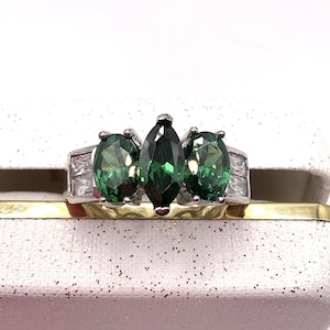 May include: A silver ring with three green gemstones. The center stone is marquise-cut, flanked by two oval-cut stones. The ring is set on a gold band and displayed on a white jewelry box.