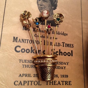 Puede incluir: Un alfiler de dedal de oro con seis alfileres decorativos, incluyendo una rosa, una mariposa, una serpiente, una flor y un camafeo, todos dispuestos en un ramo. Los alfileres están unidos al dedal con alambre dorado. El dedal está decorado con un borde de perlas.