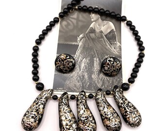 May include: A vintage jewelry set featuring a black beaded necklace with teardrop pendants and matching earrings. The pendants and earrings have a black base with gold and silver floral designs. A black and white photo is in the background.