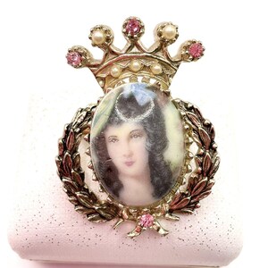 May include: Vintage gold-tone brooch featuring a portrait of a woman within an oval frame, surrounded by a laurel wreath. The brooch is topped with a crown adorned with pink rhinestones and faux pearls. A bow with a pink rhinestone is at the bottom.