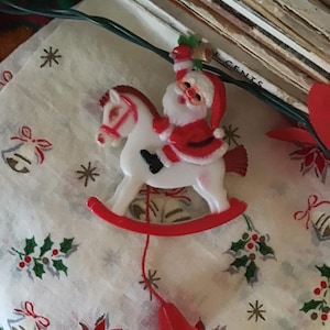 May include: A vintage Christmas ornament of Santa Claus on a white rocking horse. The ornament is red, white, and black. A red bell hangs below the horse. The ornament is on a white cloth with red and green floral patterns.