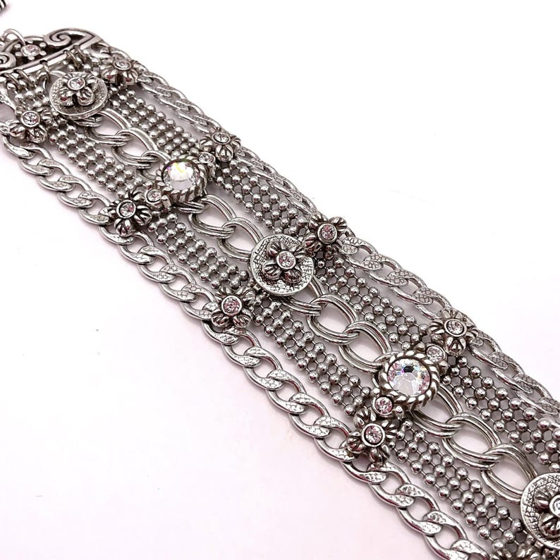 May include: Silver-toned bracelet featuring multiple strands of varying designs. The bracelet incorporates floral accents with small, pink gemstones and larger, clear crystals. The design includes chain links, beaded strands, and decorative floral elements.