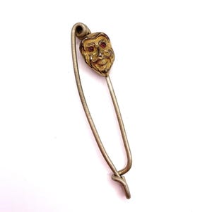 May include: Vintage safety pin with a decorative head featuring a detailed face with red accents. The pin is made of silver-toned metal and has a classic safety pin design. The face has a gold-colored finish.