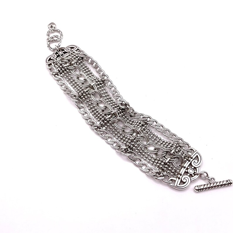 May include: Silver-toned bracelet featuring multiple chain styles and decorative elements. The bracelet has a toggle clasp and intricate detailing, showcasing a mix of chain links and beaded strands. The design is symmetrical and ornate, suitable for adding a touch of elegance.