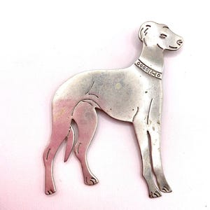 May include: A silver-toned metal brooch shaped like a standing greyhound dog. The piece features engraved lines to define the dog's muscular legs and body, along with a detailed collar around its neck. The surface has a smooth, polished finish.