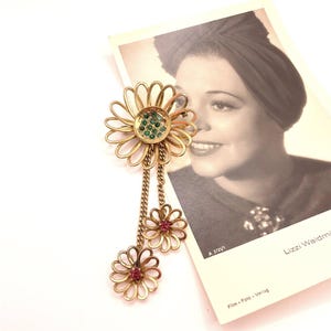 May include: Gold tone flower brooch with green and pink rhinestones. The brooch has a large flower with a center of green rhinestones and two smaller flowers with pink rhinestones hanging from the center flower.