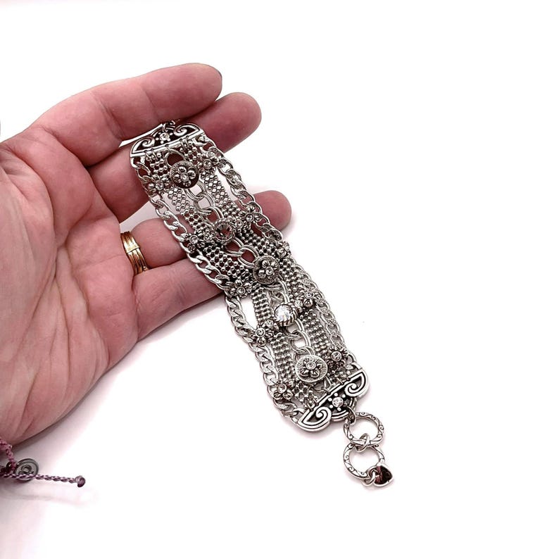 May include: Silver-toned bracelet with intricate detailing. The bracelet features multiple chains, floral accents, and clear gemstones. The design includes a series of interconnected elements, creating a textured and ornate appearance. The bracelet is held against a white background.
