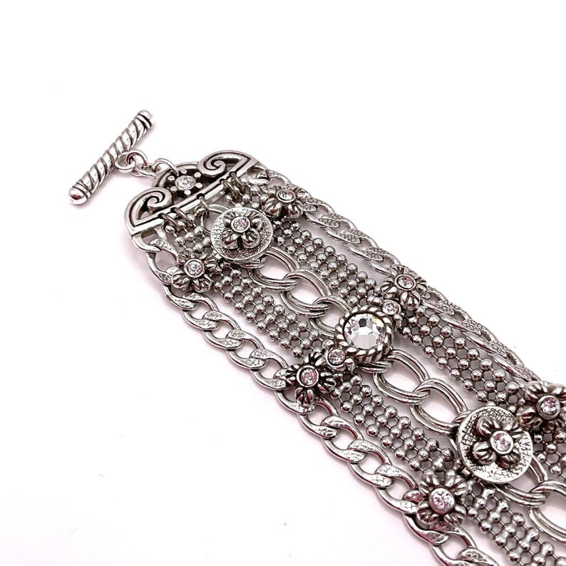 May include: Silver-toned bracelet featuring multiple strands of varying designs, including chain links, beaded rows, and floral accents. The bracelet has a toggle clasp and is adorned with clear and pink crystals.