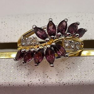 May include: A two-tone ring featuring a floral design with deep purple marquise-cut gemstones and small clear stones. The ring has a gold band and silver accents, creating a striking contrast. The gemstones are arranged to resemble petals.
