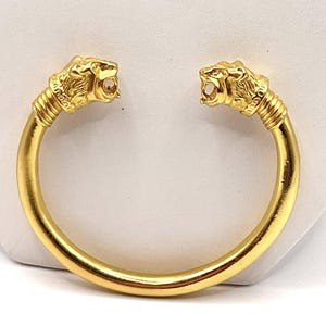 Puede incluir: Una pulsera de puño dorada con dos cabezas de león mirando hacia afuera. Las cabezas de león tienen melenas texturizadas y bocas abiertas. La pulsera tiene una banda lisa y redondeada. El diseño es audaz y llamativo.