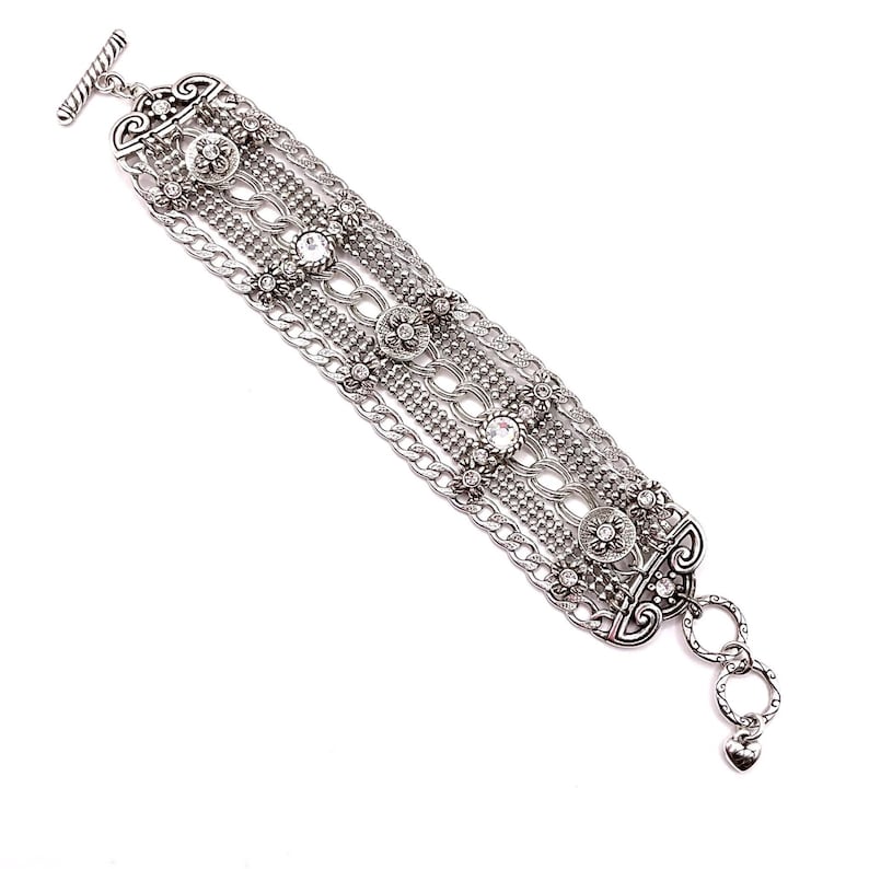 May include: Silver-toned bracelet featuring multiple strands of chain and decorative elements. The bracelet includes floral and circular designs, with clear crystal accents. A toggle clasp secures the bracelet, and a heart charm dangles from the end.