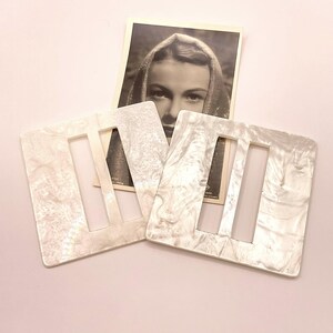 May include: Two white, square-shaped, plastic belt buckles with a rectangular cut-out in the center. The buckles have a marbled, pearlescent finish.