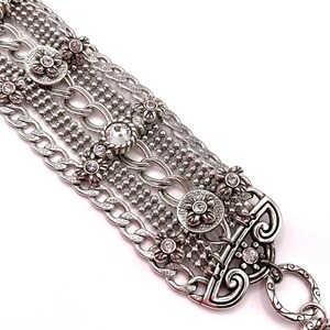 May include: Silver-toned bracelet featuring multiple chain styles, including curb and beaded chains. The bracelet is adorned with floral accents and clear and pink crystals. The clasp has an ornate design.