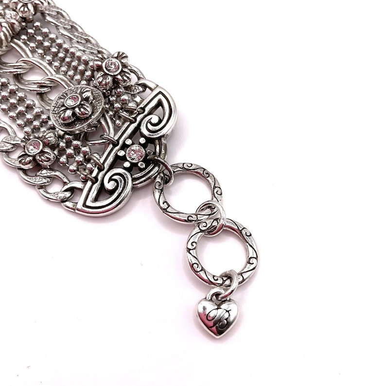 May include: Silver-toned bracelet with intricate floral and scrollwork details. The bracelet features multiple chain strands, decorative flower accents with clear crystals, and a heart-shaped charm. The design includes a series of connecting rings.
